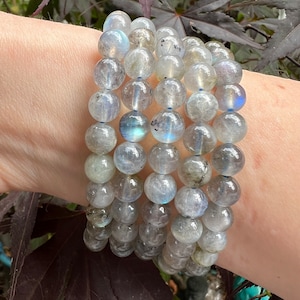 May include: Four stacked bracelets made of iridescent grey labradorite beads. The bracelets are made of round beads and are stacked on a person's wrist.