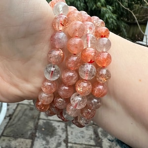 Puede incluir: Un brazalete de múltiples hilos hecho de cuentas redondas de piedra de sol translúcida de color naranja-rojo. Las cuentas tienen un acabado brillante y metálico y están enhebradas en hilo rojo.