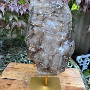 May include: A large, smoky quartz crystal cluster mounted on a gold-colored metal stand. The crystal has a layered, geometric structure with varying shades of brown and gray. The stand is square with a central post.