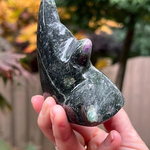 May include: A dark green, polished stone sculpture with a smooth, curved shape. The stone has a marbled appearance with flecks of lighter green and a small, round, pinkish-purple accent. The sculpture is held in a hand.