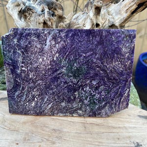 Puede incluir: Un bloque rectangular de piedra de charoíta, que muestra una rica mezcla de tonos púrpuras con patrones arremolinados y toques de verde. La superficie de la piedra tiene un aspecto texturizado, con estructuras cristalinas visibles. Ideal para coleccionistas y artesanos.