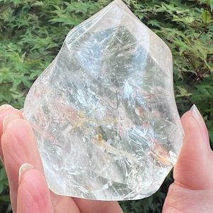 May include: A clear, polished quartz crystal point with a pointed top and a wide, rounded base. The stone features internal fractures, natural inclusions, and a transparent, glass-like appearance with subtle golden-brown mineral deposits inside.