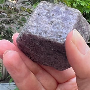 Könnte beinhalten: Ein rohes, sechseckiges Kristall-Exemplar mit einer rauen, strukturierten Oberfläche in staubigen Violett- und Grautönen. Das Mineral zeigt eine natürliche, unpolierte Kristallstruktur mit sichtbaren Facetten und erdigen, mineralischen Einschlüssen.