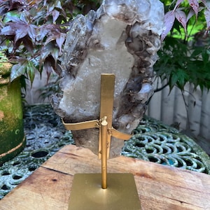 May include: A large, raw crystal cluster with a mix of clear and smoky quartz, mounted on a gold-colored metal stand. The crystal is irregularly shaped and has a rough texture. The stand has a square base and a vertical support with curved arms holding the crystal.
