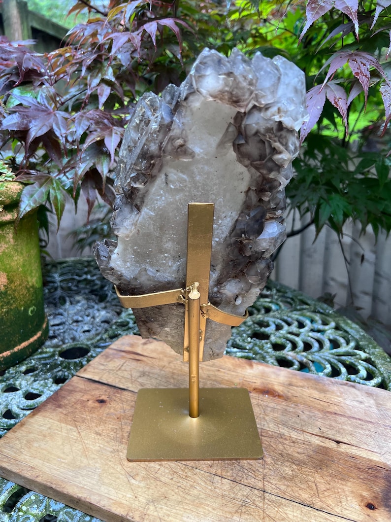 May include: A large, raw crystal cluster with a mix of clear and smoky quartz, mounted on a gold-colored metal stand. The crystal is irregularly shaped and has a rough texture. The stand has a square base and a vertical support with curved arms holding the crystal.