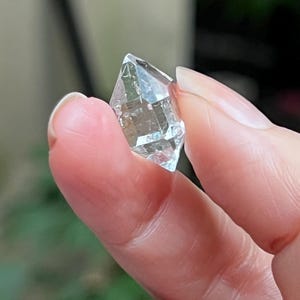 Peut inclure: Un petit cristal de quartz transparent à double terminaison, doté d'une structure hexagonale et d'extrémités pointues. Le cristal présente des inclusions internes visibles et des facettes qui reflètent la lumière, révélant une forme naturelle, géométrique et prismatique.