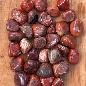 Può includere: Una collezione di pietre di diaspro rosso lucido con segni marrone scuro e grigi. Le pietre variano per dimensioni e forma, con un aspetto liscio e arrotondato. Il diaspro è esposto su una superficie di legno marrone chiaro.