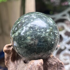 Può includere: Una pietra lucida di forma sferica con una base color verde bosco profondo, caratterizzata da intricate inclusioni maculate bianche e verde chiaro su tutta la superficie liscia e riflettente, appoggiata su un pezzo di legno naturale e strutturato.