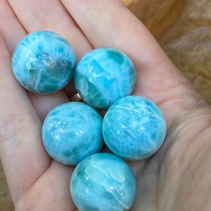 May include: Five polished, round larimar stones in varying shades of blue and white are held in an open hand. The stones have a marbled appearance, with swirls and patterns. The overall effect is a collection of smooth, ocean-toned spheres.