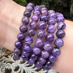 Puede incluir: Una pila de cinco pulseras de piedras preciosas de color púrpura y plateado. Cada pulsera está hecha de cuentas redondas con un acabado brillante e iridiscente.