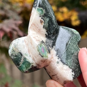 May include: A star-shaped stone sculpture in shades of green, white, and black. The polished stone is held in a hand, with a blurred backdrop of autumnal leaves. The sculpture's form is clearly defined, showcasing its unique shape.
