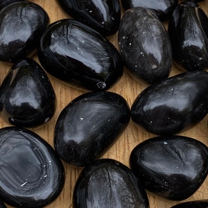 Könnte beinhalten: Eine Sammlung glatter, schwarzer Obsidian-Trommelsteine. Die Steine sind poliert und haben eine glänzende Oberfläche.