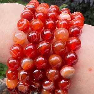 May include: A bracelet made of carnelian beads. The beads are a deep orange colour with white stripes. The bracelet is on a person's wrist.