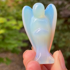 May include: A small, white opal angel figurine with iridescent wings. The angel is carved in a standing pose with its hands clasped in front of its chest.