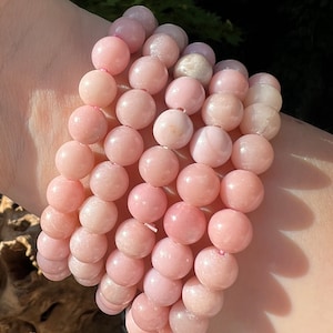 Könnte beinhalten: Ein Stapel rosafarbener Opal-Armbänder. Jedes Armband besteht aus runden, glatten Perlen.