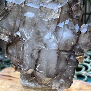 May include: A cluster of smoky quartz crystals, showcasing a range of translucent and opaque sections. The crystals are mounted on a gold-colored stand, with visible facets and natural formations. The overall color palette is a mix of gray, brown, and clear.