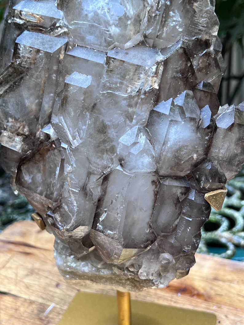 May include: A cluster of smoky quartz crystals, showcasing a range of translucent and opaque sections. The crystals are mounted on a gold-colored stand, with visible facets and natural formations. The overall color palette is a mix of gray, brown, and clear.