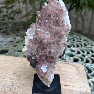 Pode incluir: Um aglomerado de cristais naturais em tons claros, brancos e castanho-avermelhados. Os cristais são montados em um suporte de metal preto. A formação áspera e texturizada é exibida em um fundo verde desfocado.