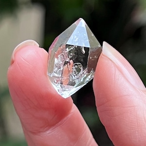 Peut inclure: Un cristal de quartz transparent à double terminaison, de forme hexagonale avec des extrémités pointues. La pierre présente des inclusions internes, des fractures et des facettes naturelles qui reflètent la lumière, créant un aspect transparent.