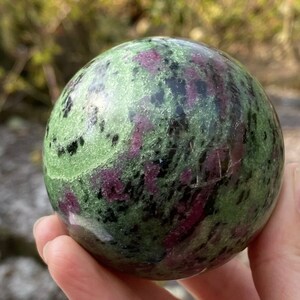 May include: A green, black and pink gemstone sphere. The sphere is smooth and polished, with a variety of colours and patterns.
