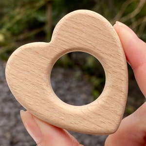 May include: A light-colored, smooth wooden heart-shaped object featuring a circular cutout in the center. The natural wood grain is visible across the surface of the heart, which has rounded edges and a solid, flat construction throughout the piece.