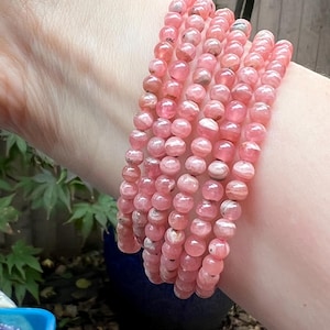 Könnte beinhalten: Mehrere rosa Edelsteinarmbänder sind auf einem Handgelenk gestapelt. Die Armbänder bestehen aus runden, polierten Perlen mit transluzentem Aussehen. Die Perlen haben eine Reihe von Rosatönen, wobei einige hellere und dunklere Variationen aufweisen. Die Armbänder sind ein natürliches, erdiges Accessoire.