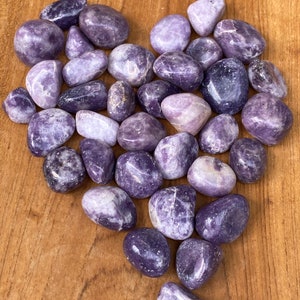 May include: A collection of polished, tumbled purple stones arranged in a heart shape. The stones vary in shades of lavender and deep violet, with smooth, rounded surfaces. They are displayed on a light brown wooden surface.