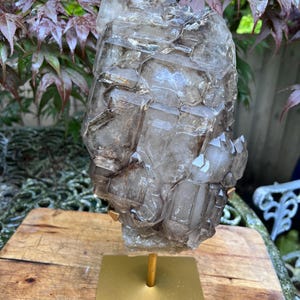May include: A large, translucent smoky quartz crystal cluster, mounted on a gold-colored metal stand. The crystal has a complex, geometric structure with varying shades of brown and gray. The stand has a square base and a central post.