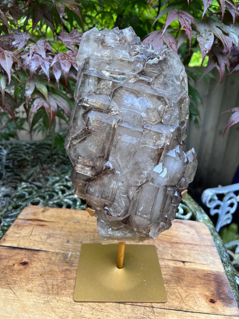May include: A large, translucent smoky quartz crystal cluster, mounted on a gold-colored metal stand. The crystal has a complex, geometric structure with varying shades of brown and gray. The stand has a square base and a central post.