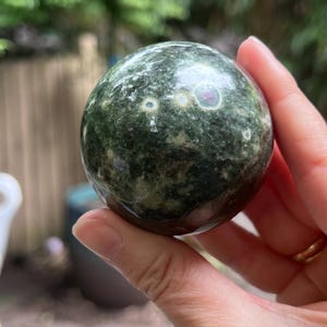 May include: A polished, spherical stone featuring a deep forest green base color with mottled textures and distinct circular inclusions in shades of white, cream, and a small accent of magenta-pink, showcasing a smooth, reflective surface.