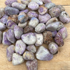 May include: A collection of tumbled purple and white stones with a variety of patterns and textures. The stones are smooth and polished, and they are arranged in a pile on a wooden surface.