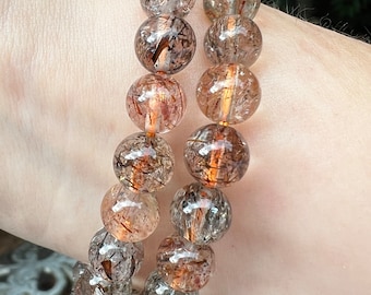 May include: Two beaded bracelets with clear, round beads. Each bead has brown and orange inclusions, creating a unique pattern. The bracelets are worn on a wrist, showcasing the natural colors and textures of the gemstones.