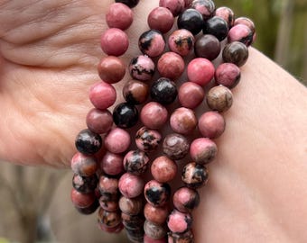 May include: A stack of pink and black gemstone bracelets. The bracelets are made of round beads and are stacked on a person's wrist.