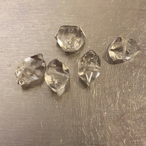 Puede incluir: Una colección de seis cuentas de cristal transparentes y facetadas. Las cuentas tienen varias formas, incluyendo hexagonal y de lágrima, y están dispuestas sobre una superficie plateada. Los cristales tienen un aspecto brillante y transparente.