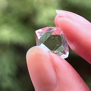 Peut inclure: Une petite pierre précieuse transparente et facettée de forme géométrique. La pierre présente de multiples surfaces plates et polies qui reflètent la lumière, créant des bords nets et un aspect translucide. Le cristal est tenu entre deux doigts.