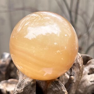 May include: A smooth, polished, orange calcite sphere. The sphere is sitting on a piece of driftwood.
