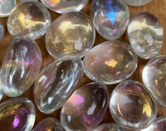 May include: A cluster of iridescent, oval-shaped stones with a polished surface. The stones reflect a range of colours, including gold, purple, and blue, creating a shimmering effect. The stones are scattered on a wooden surface.