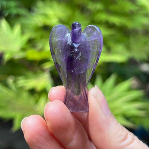May include: A small, carved amethyst figurine in the shape of an angel with translucent purple hues. The crystal sculpture features defined wings, a rounded head, and a tapered body, showcasing natural variations in the deep violet stone color.