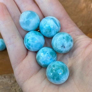 May include: A hand holds six polished, round larimar gemstones. The stones vary in shades of blue and white, with some displaying a turquoise hue. The spherical stones are smooth and appear to be natural, with unique patterns.
