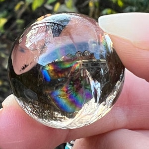 Pode incluir: Uma conta esférica de vidro transparente com uma inclusão interna iridescente nas cores do arco-íris. A superfície da conta reflete o ambiente, incluindo árvores e um edifício, criando uma vista panorâmica em miniatura distorcida.