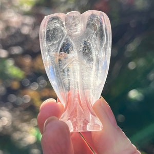 May include: A clear quartz angel figurine with outstretched wings and a detailed carved design. The angel is held in a hand against a blurred green and brown background. The figurine has a translucent quality.