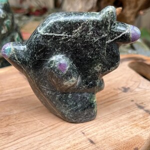 May include: A dark green stone carving of a stylized animal figure with a pointed ear and a raised arm. The sculpture has a polished finish with purple accents on the ear and body. The carving is displayed on a wooden surface.