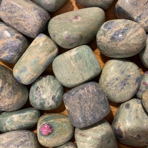 May include: A collection of green, blue and grey tumbled stones with red ruby inclusions. The stones are smooth and polished, and they have a variety of shapes and sizes.