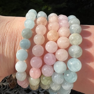 May include: A stack of five bracelets made of round, smooth, pastel-coloured gemstone beads. The bracelets are layered on a person's wrist, with the beads in shades of light blue, pink, and green.