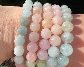 May include: A stack of five bracelets made of round, smooth, pastel-colored gemstone beads. The bracelets are layered on a person's wrist, with the beads in shades of light blue, pink, and green.