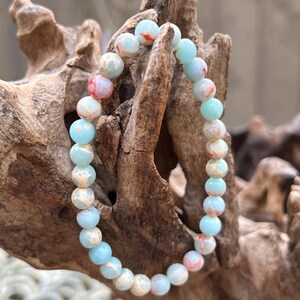 May include: A light blue and white bracelet with red and gold accents. The bracelet is made of small round beads and is strung on an elastic cord.