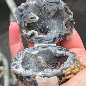 Puede incluir: Una geoda dividida con un exterior veteado gris y blanco, que revela un interior cristalino brillante. La geoda se mantiene abierta, mostrando el centro ahuecado lleno de pequeños cristales y una base oscura y terrosa. La cáscara exterior tiene una apariencia rugosa y texturizada.