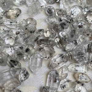 Peut inclure: Une collection de pointes de quartz clair avec des inclusions noires. Les pointes sont de tailles et de formes variées et sont dispersées sur un fond de tissu blanc.