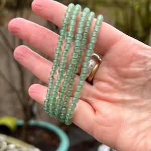 Pode incluir: Cinco pulseiras de contas verdes empilhadas. As pulseiras são feitas de pequenas contas redondas de um tom verde claro.