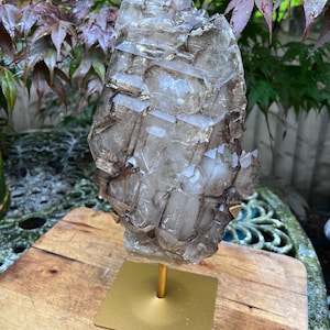 May include: A large, smoky quartz crystal cluster mounted on a gold-colored metal stand. The crystal has a complex, geometric structure with multiple facets and a translucent appearance. The stand has a square base and a slender, vertical support.